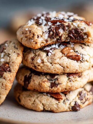 A stack of homemade forgotten cookies with melted chocolate chunks, toasted pecans, and flaky sea salt on top, sitting on a rustic plate in warm natural light.