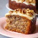 Homemade Banana Walnut Cake Recipe slice on a pink plate, topped with creamy frosting and chopped walnuts, with the rest of the cake blurred in the background.