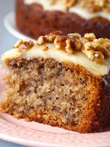 Homemade Banana Walnut Cake Recipe slice on a pink plate, topped with creamy frosting and chopped walnuts, with the rest of the cake blurred in the background.