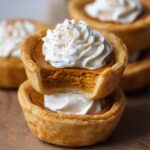 Homemade mini pumpkin pies with flaky golden crusts, creamy pumpkin filling, and whipped cream on top, one showing a bite for a cozy, rustic look.
