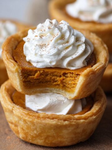 Homemade mini pumpkin pies with flaky golden crusts, creamy pumpkin filling, and whipped cream on top, one showing a bite for a cozy, rustic look.