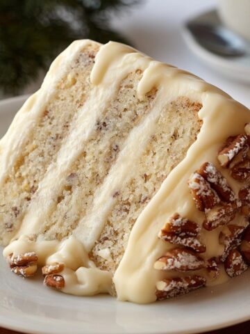 Homemade Snowy Bavarian Bliss Cake slice with soft vanilla layers, creamy white frosting, and pecans on a white plate, captured in warm natural light.