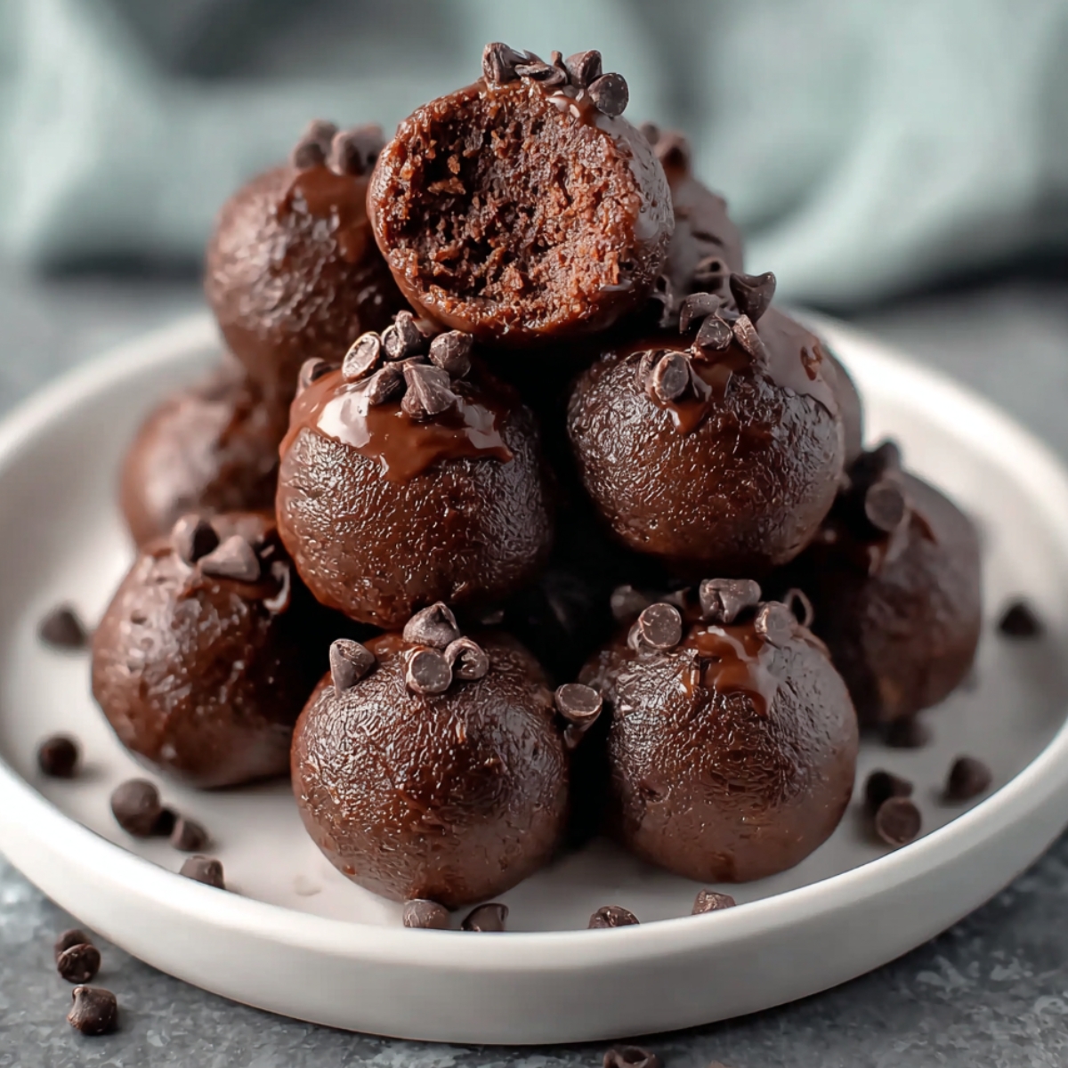 Homemade no-bake brownie bites stacked on a small ceramic plate, topped with melted chocolate and cacao nibs, with one bite showing the fudgy, rich texture inside.