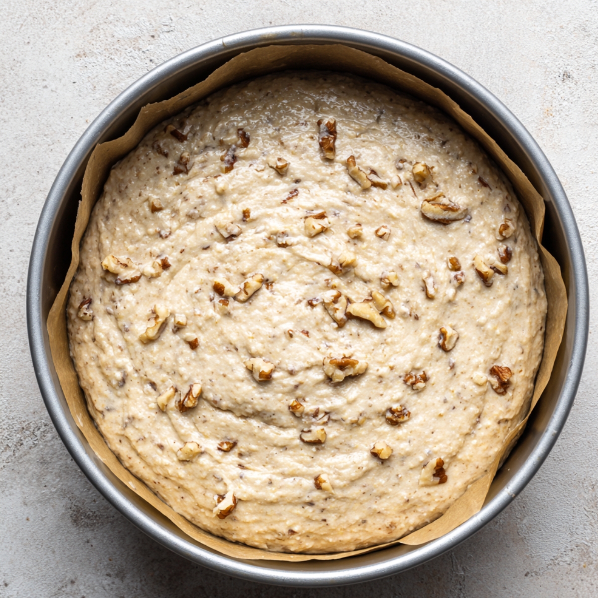 Top-down view of banana walnut cake batter in a round parchment-lined pan, light golden with walnut pieces on top, under natural light.
