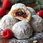 Close-up of homemade Raspberry Filled Almond Snowball Cookies coated in powdered sugar, one cut open showing raspberry filling, with fresh raspberries and soft natural light on a wooden surface.
