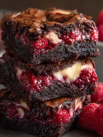 Raspberry Swirl Brownies with a fudgy chocolate texture, bright red raspberries, and creamy swirls, sitting on a gray surface beside fresh raspberries and a blurred bowl in the background.