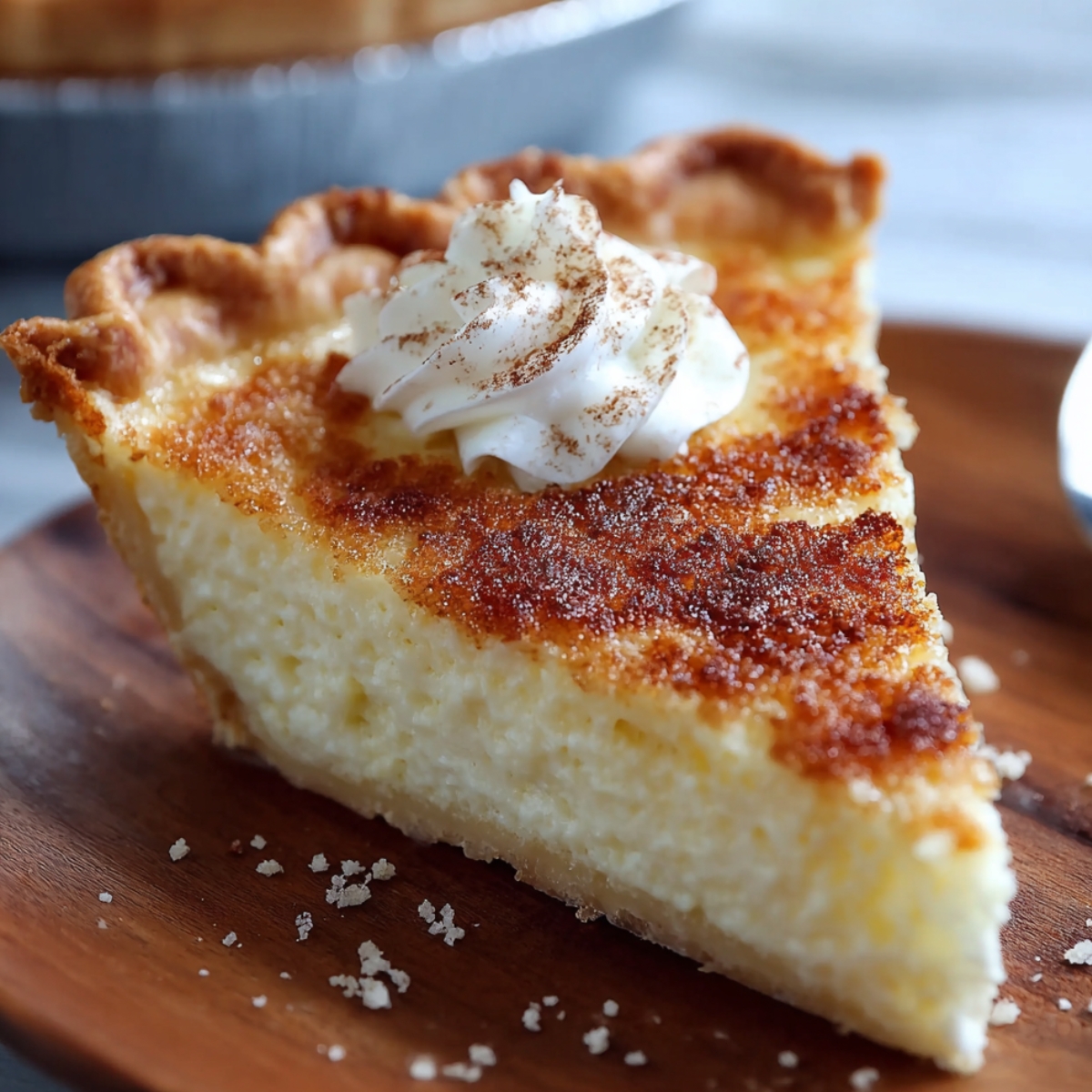 Homemade sugar cream pie slice with golden crust, creamy filling, caramelized top, and whipped cream dusted with cinnamon on a wooden plate.