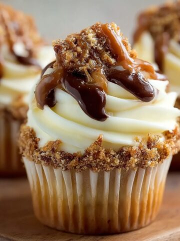 Close-up of homemade Toffee Crunch Cupcakes with vanilla frosting, caramel and chocolate drizzle, and crushed toffee crumbs on a wooden board in warm natural light.
