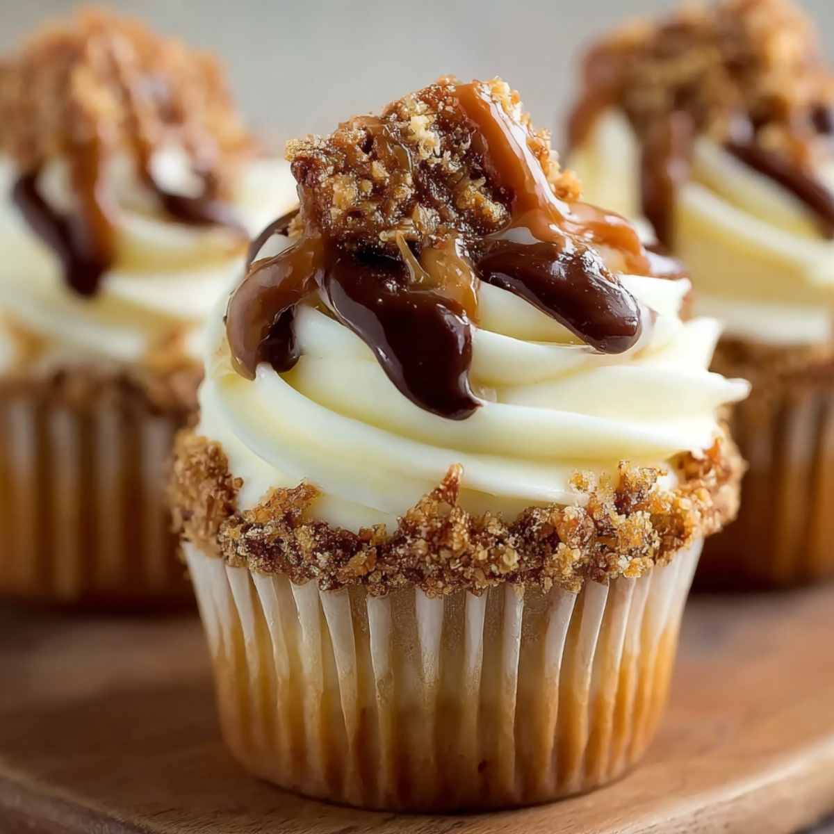 Close-up of homemade Toffee Crunch Cupcakes with vanilla frosting, caramel and chocolate drizzle, and crushed toffee crumbs on a wooden board in warm natural light.