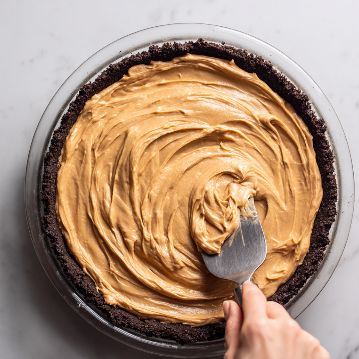 Hand spreading creamy peanut butter filling over a chocolate cookie crust in a glass pie dish on a marble countertop.