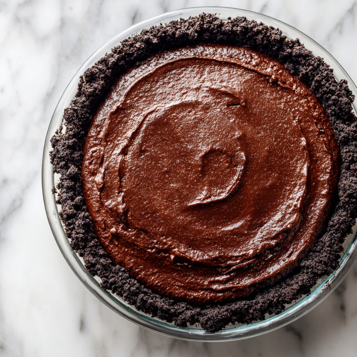 A homemade Mississippi Mud Pie with a smooth, rich chocolate filling spread inside a dark Oreo cookie crust in a glass pie dish on a marble countertop.