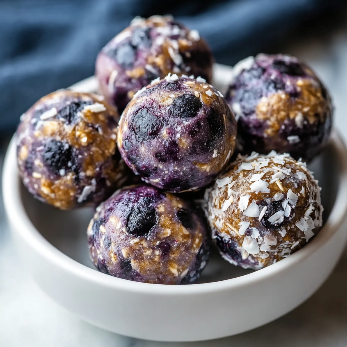 Blueberry Protein Balls stacked in a small bowl, showing their deep purple color, visible oats, and soft, chewy texture.