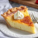 A slice of homemade buttermilk pie with a golden caramelized top, flaky crust, and a dollop of whipped cream on a white plate with a fork.