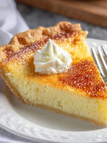 A slice of homemade buttermilk pie with a golden caramelized top, flaky crust, and a dollop of whipped cream on a white plate with a fork.