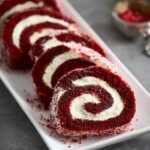 Christmas Red Velvet Cake Roll sliced on a dark serving board, showing its vibrant red sponge and thick swirl of creamy white filling, lightly dusted with powdered sugar.