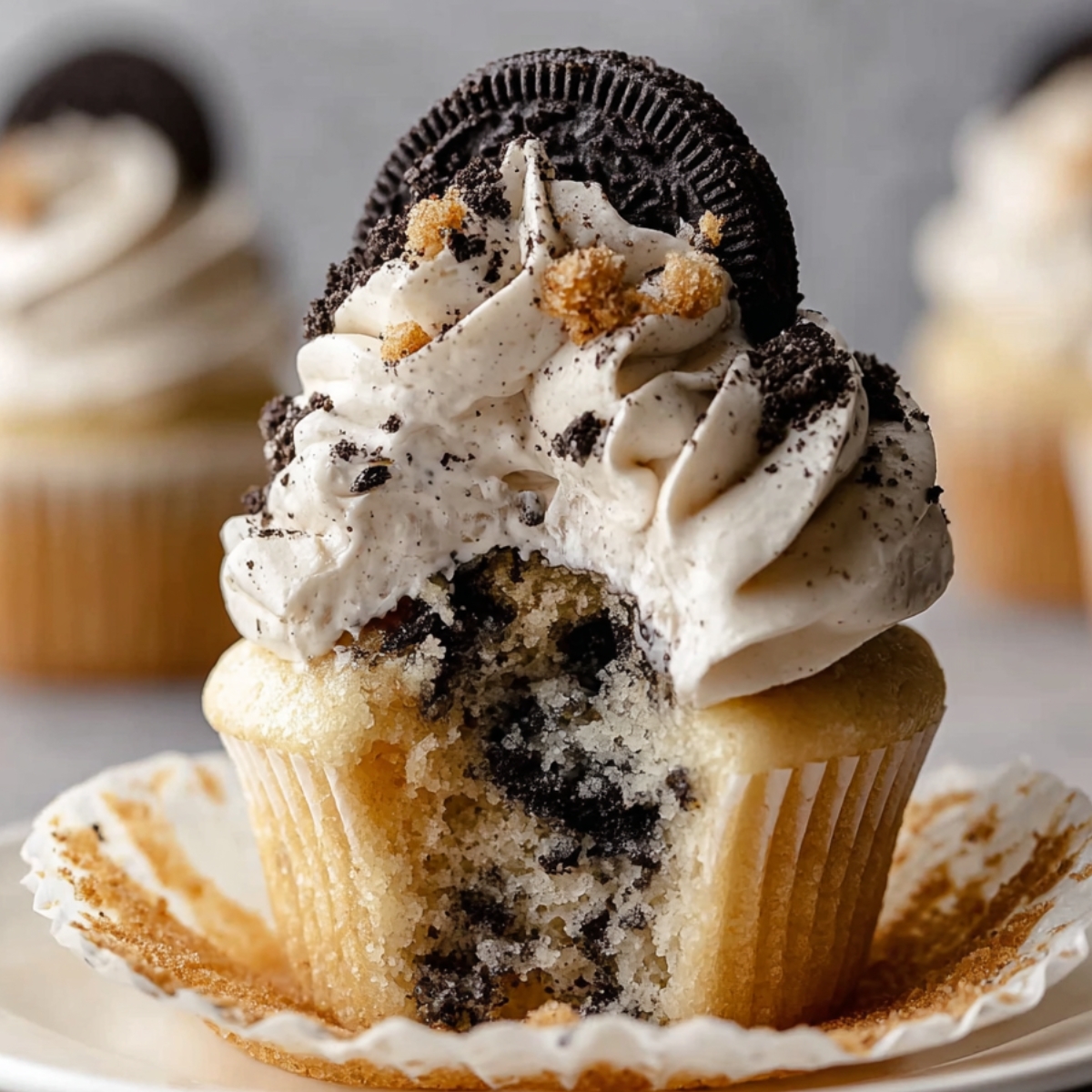Cookies and Cream Cupcakes with creamy vanilla frosting, crushed cookie bits, and a whole Oreo on top, with the paper liner peeled back and a soft bite revealing chunks of cookies inside.