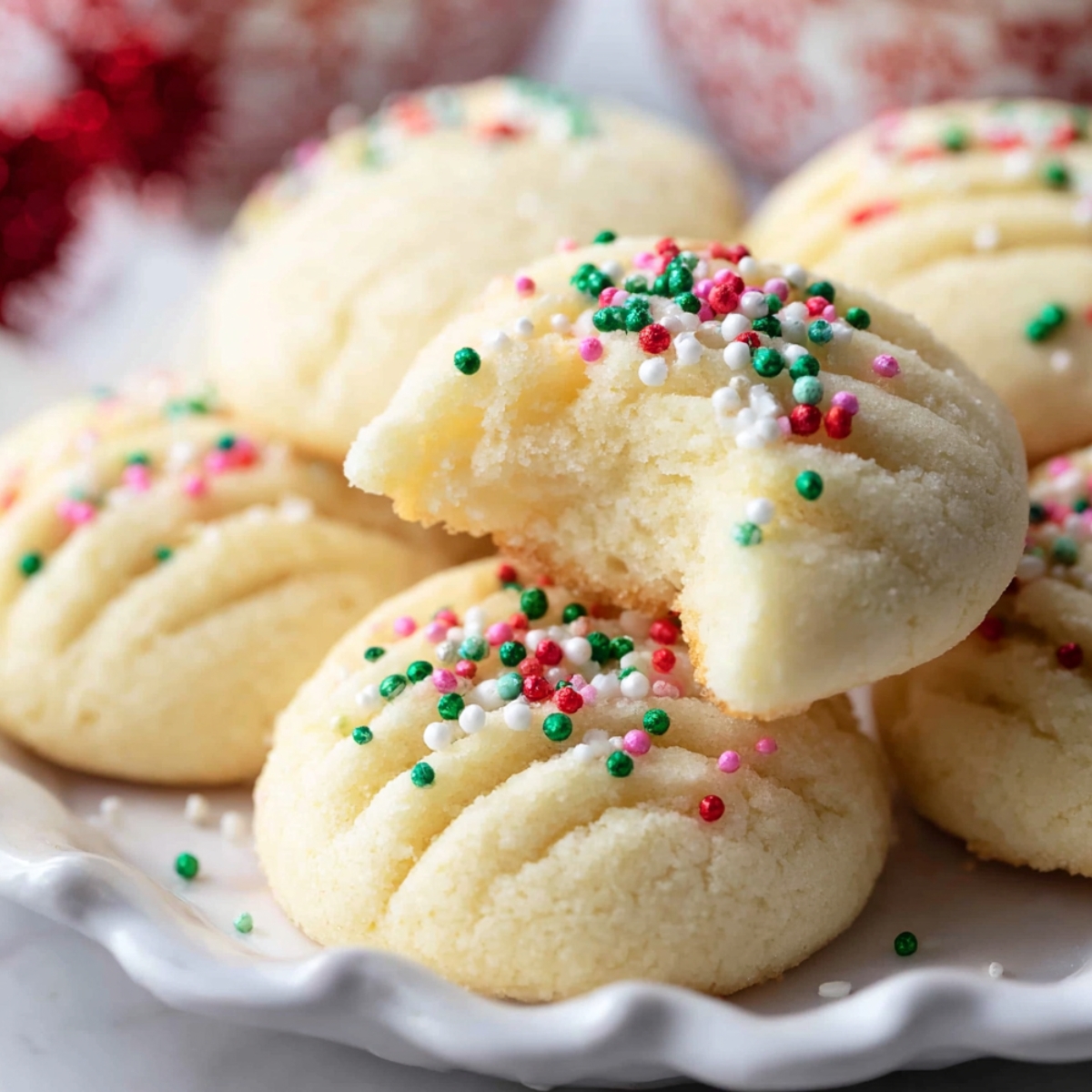 Plate of soft homemade whipped shortbread cookies topped with red, green, and white sprinkles, with one cookie showing a bite taken out to reveal its tender, crumbly texture.