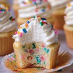 Funfetti Vanilla Cupcakes with a bite taken out, showing a soft, fluffy vanilla crumb filled with colorful rainbow sprinkles, topped with creamy white buttercream frosting and extra sprinkles, with other frosted cupcakes blurred in the background.