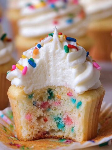 Funfetti Vanilla Cupcakes with a bite taken out, showing a soft, fluffy vanilla crumb filled with colorful rainbow sprinkles, topped with creamy white buttercream frosting and extra sprinkles, with other frosted cupcakes blurred in the background.