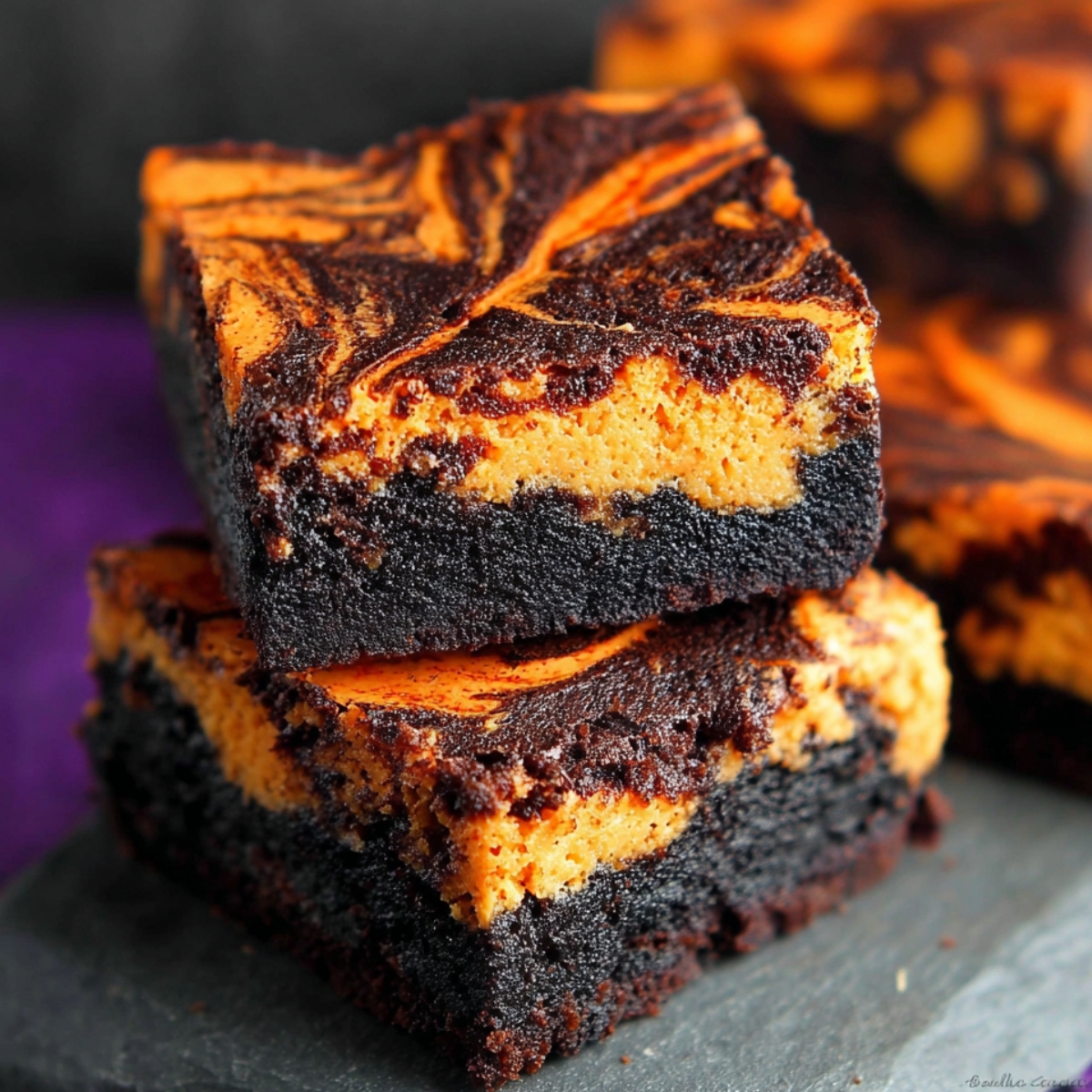 Halloween Swirl Cream Cheese Brownies stacked on a dark slate board, showing deep chocolate layers and marbled orange cheesecake swirls with a fudgy, moist texture.
