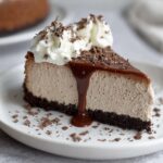 Homemade Baileys Cheesecake topped with whipped cream, chocolate shavings, and warm chocolate sauce dripping down the side on a white plate.