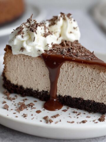 Homemade Baileys Cheesecake topped with whipped cream, chocolate shavings, and warm chocolate sauce dripping down the side on a white plate.