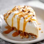 Homemade Caramel Cream Pie on a beige plate, topped with whipped cream, golden caramel drizzle, and crushed candy pieces over a crumbly graham cracker crust.