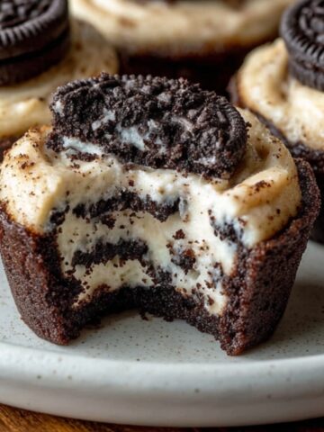 Homemade Oreo Cheesecake Cookie Cups with creamy cheesecake filling, Oreo layers, and a bite taken out on a white plate.