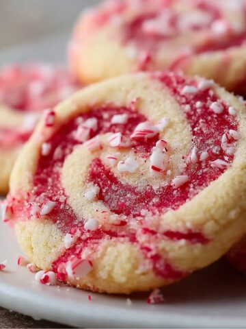Homemade peppermint swirl cookies with red and white spiral dough, topped with crushed candy cane pieces and a light sugar sparkle, resting on a white plate — soft, buttery, and festive for the holidays.