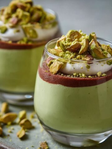 Homemade pistachio mousse in small glass cups topped with chocolate ganache, whipped cream, and chopped pistachios.