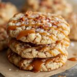 Stack of homemade salted caramel crunch cookies drizzled with caramel and sprinkled with flaky sea salt on a piece of parchment paper.