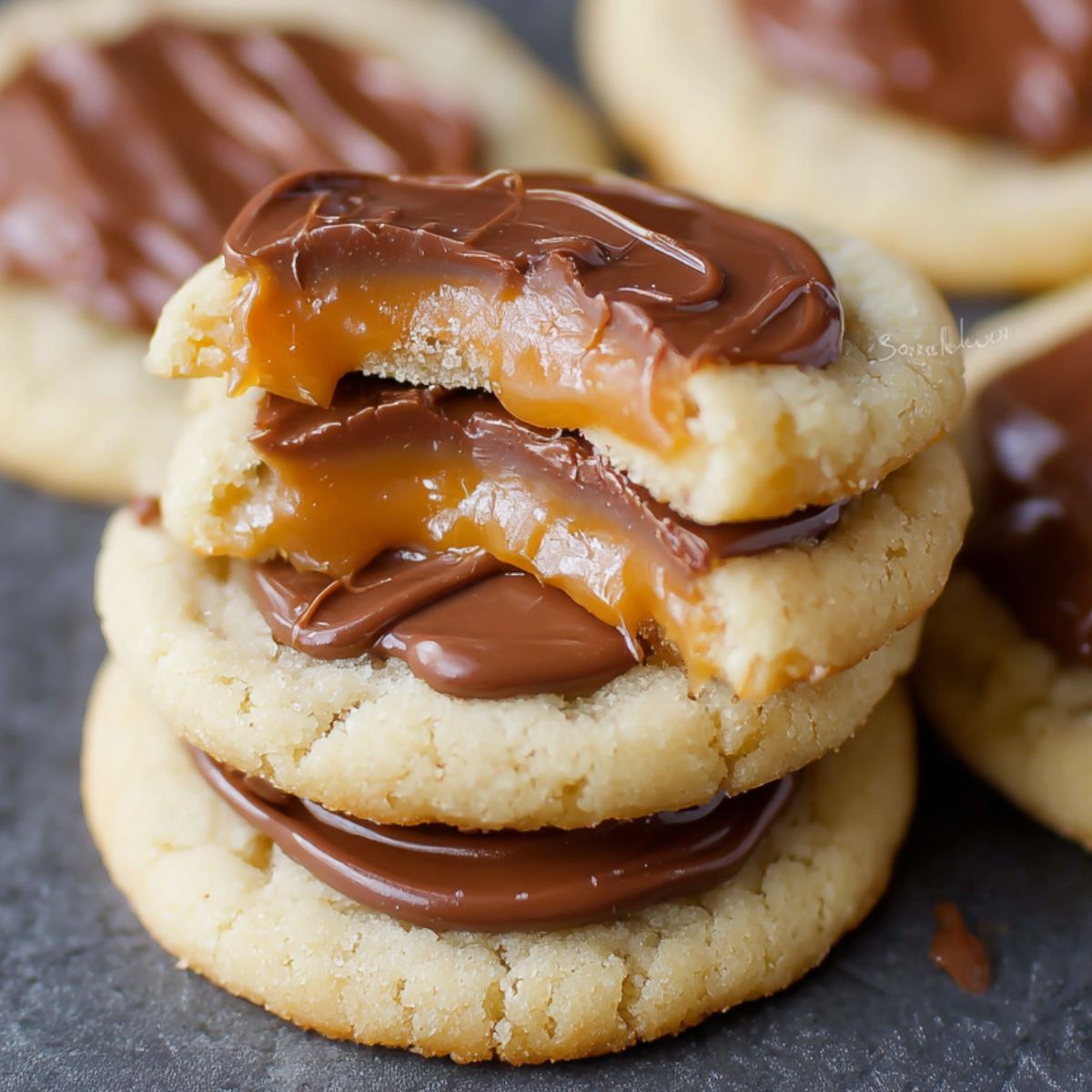 Homemade Twix Cookies topped with melted chocolate and gooey caramel, with one cookie showing a bite revealing soft shortbread, creamy chocolate, and dripping caramel.