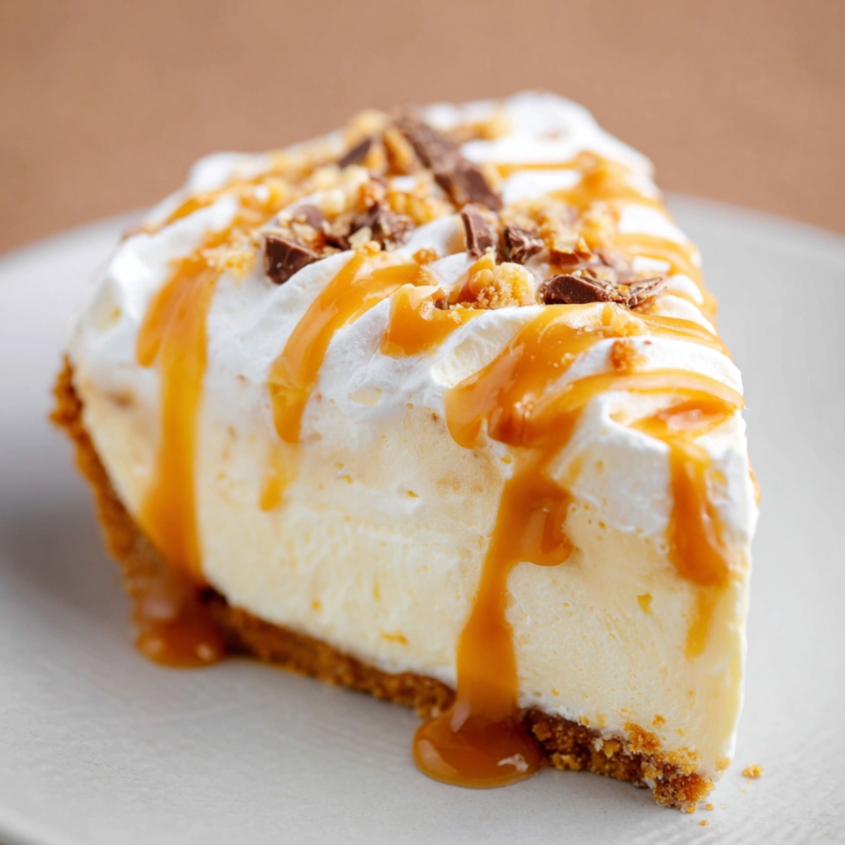 A slice of homemade caramel cream pie on a white plate, topped with whipped cream, drizzled caramel sauce, and sprinkled chopped nuts over a golden graham cracker crust.