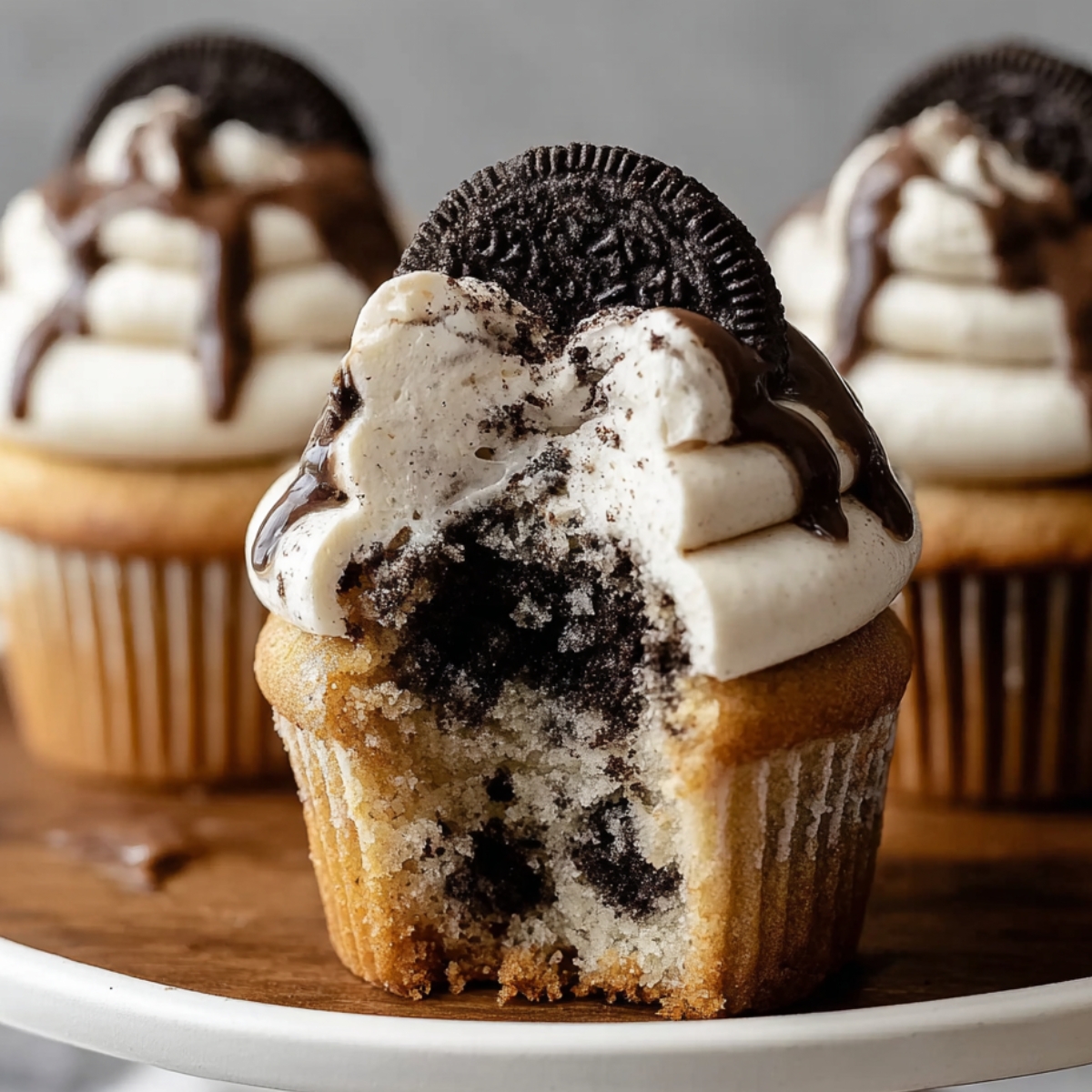 Homemade cookies and cream cupcake topped with vanilla frosting, a drizzle of chocolate syrup, and a whole Oreo, with a soft bite showing crushed cookies baked inside.