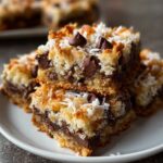 Stacked homemade Magic Cookie Bars on a white plate, showing layers of chocolate chips, toasted coconut, and a golden graham crust, with a rustic, real-kitchen look.