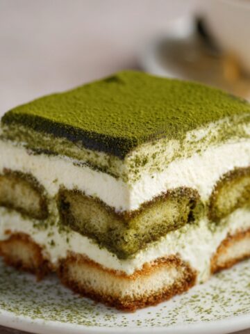 Matcha Tiramisu on a white plate, showing layers of creamy mascarpone and matcha-soaked ladyfingers, topped with a thick dusting of matcha powder.