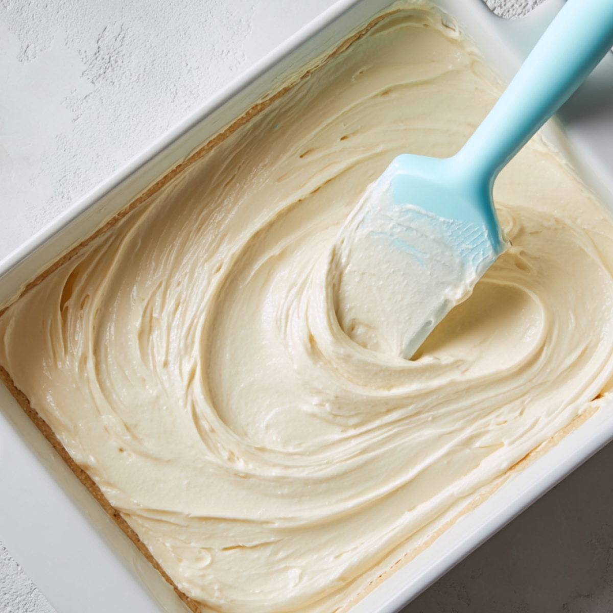 Cheesecake filling being spread over a crust in a baking dish with a blue silicone spatula.