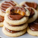 Twix Cookies topped with melted chocolate and gooey caramel, with one cookie showing a bite revealing soft shortbread, creamy chocolate, and dripping caramel.