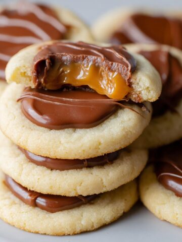 Twix Cookies topped with melted chocolate and gooey caramel, with one cookie showing a bite revealing soft shortbread, creamy chocolate, and dripping caramel.