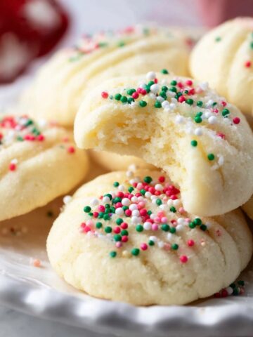 Whipped Shortbread Cookies on a plate, topped with red, green, and white sprinkles, with one cookie bitten to show its soft, crumbly inside.