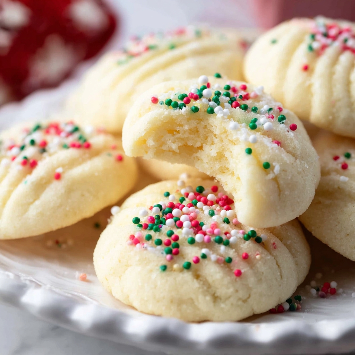 Whipped Shortbread Cookies on a plate, topped with red, green, and white sprinkles, with one cookie bitten to show its soft, crumbly inside.