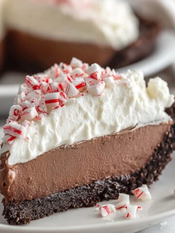 A slice of homemade chocolate peppermint mousse pie with a crumbly chocolate cookie crust, creamy chocolate mousse, fluffy whipped topping, and crushed peppermint pieces, served on a white plate.