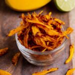 Bowl of homemade dehydrated mango slices on a marble countertop, with a halved lime in the background.