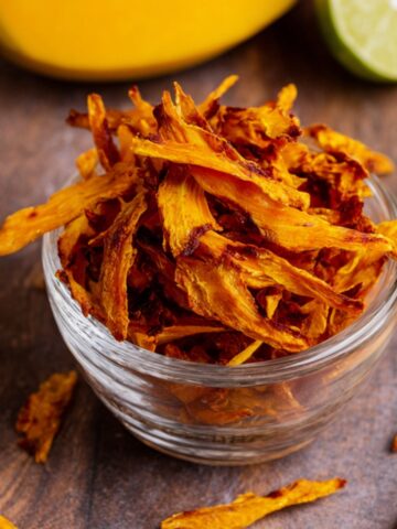 Bowl of homemade dehydrated mango slices on a marble countertop, with a halved lime in the background.