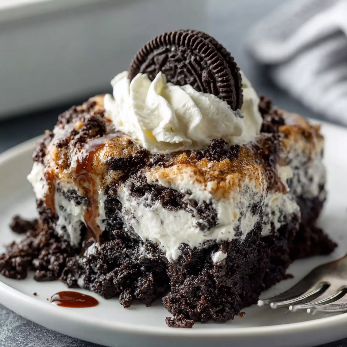 Easy Oreo Dump Cake Recipe 10 A homemade slice of Oreo Dump Cake topped with whipped cream, an Oreo cookie, and a drizzle of caramel, sitting on a rustic plate with a fork beside it.
