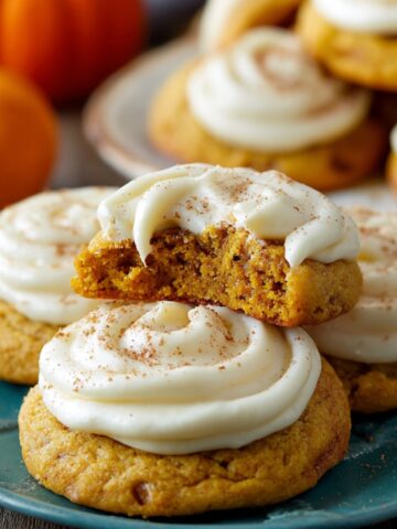 Frosted Pumpkin Cookies topped with creamy white icing, with a bitten cookie on top showing its soft, moist pumpkin center.