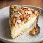 Homemade Bee Sting Cake with creamy filling and caramelized sliced almonds sits on a speckled plate beside a fork.