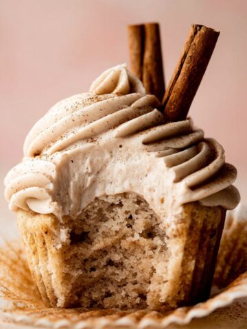 Homemade Chai Latte Cupcakes with swirled cinnamon buttercream and two cinnamon sticks on top, with a bite taken out to reveal the spiced interior.