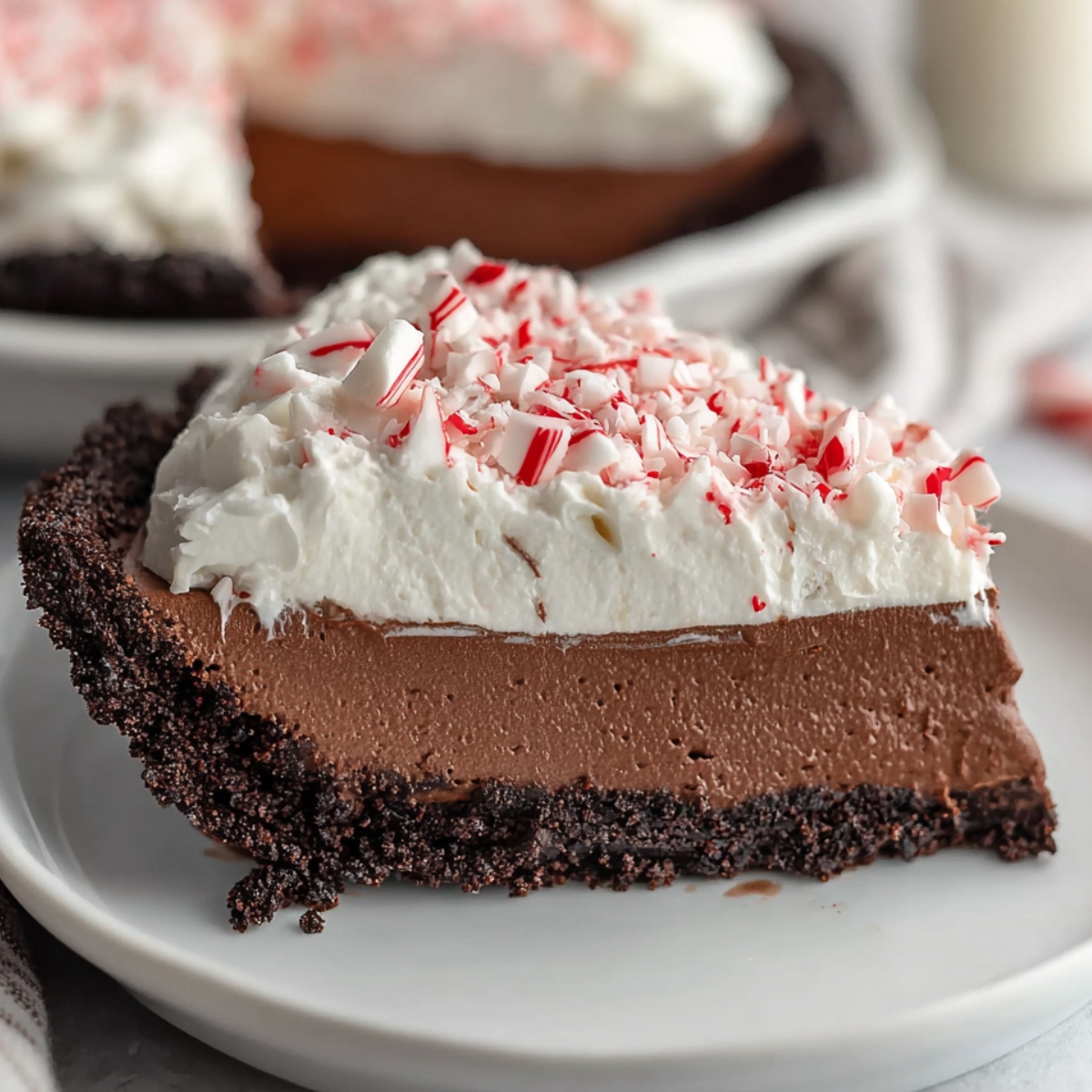 A slice of homemade chocolate peppermint mousse pie with a crumbly chocolate cookie crust, creamy chocolate mousse filling, fluffy whipped topping, and crushed peppermint pieces on top, served on a white plate.