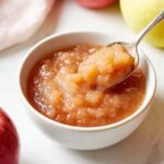 Bowl of homemade crockpot applesauce with a spoon lifting a chunky scoop, surrounded by fresh apples on a bright kitchen counter.